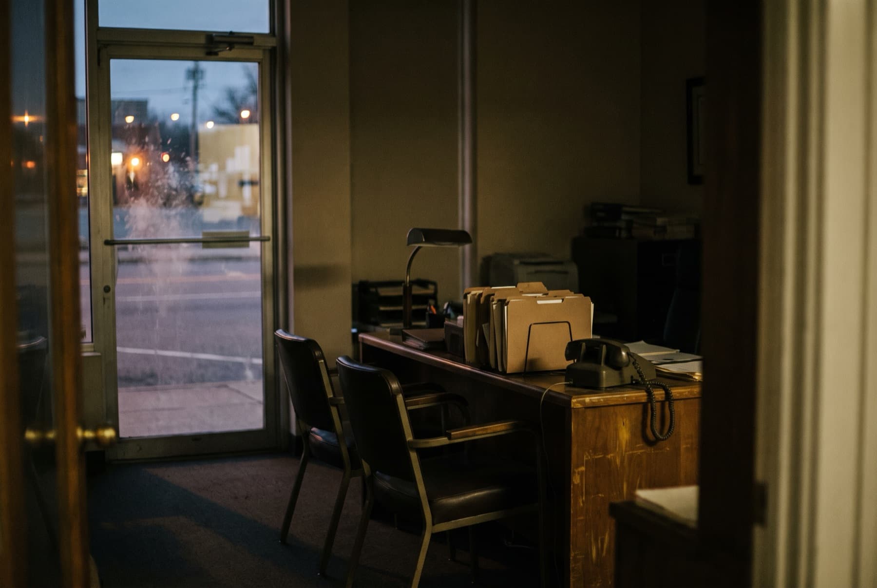 An empty, unlit small law office seen through a glass door at dusk: chairs pushed in