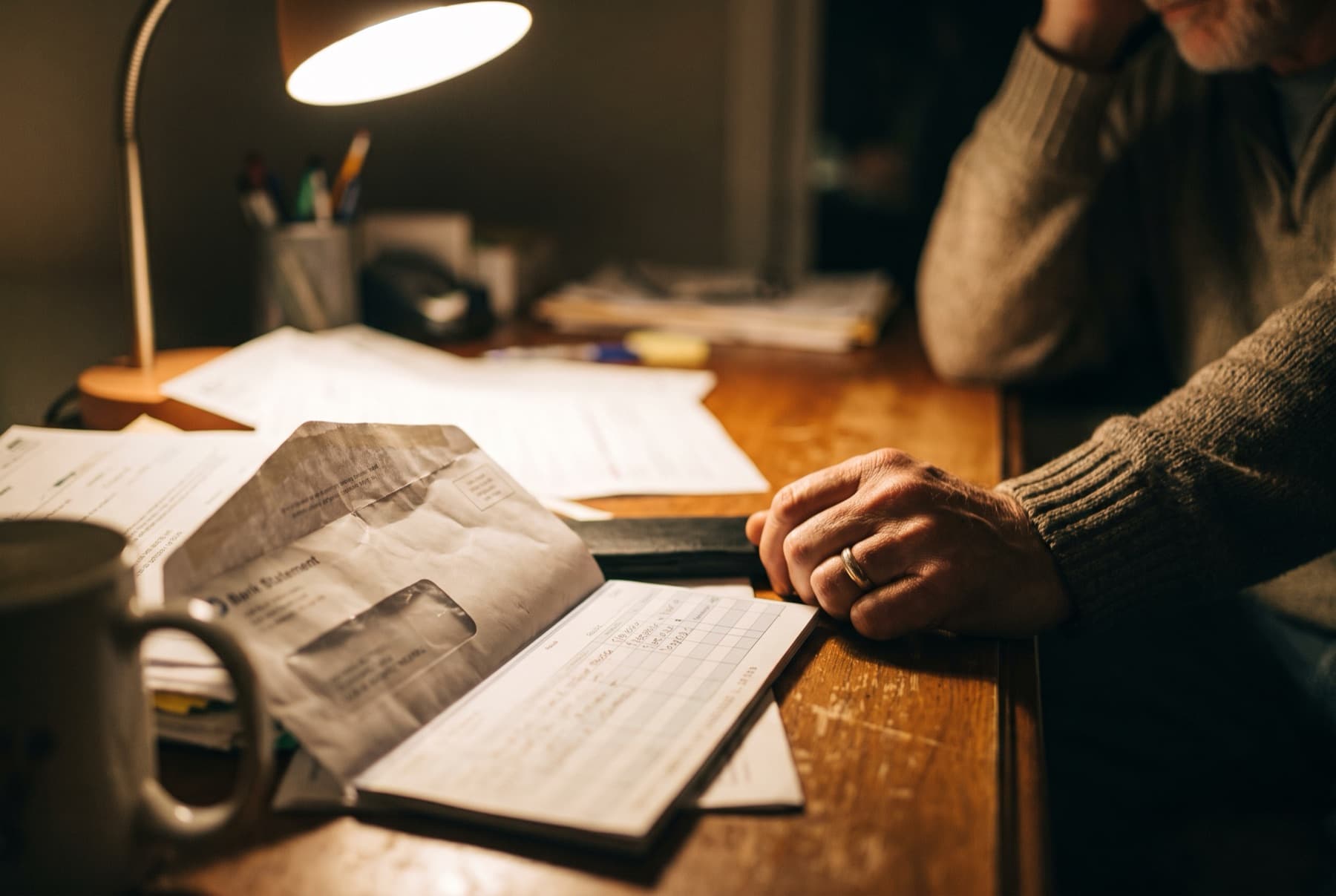 An open bank statement envelope and a checkbook register on a desk at night lit by a single lamp